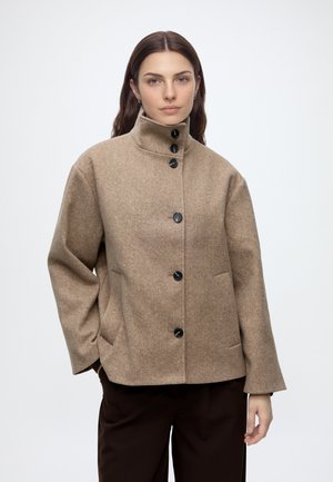 Woman wearing a beige high-collar buttoned coat and dark pants against a plain light background, posing with one hand in pocket.