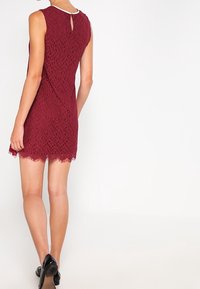 Robe en dentelle bordeaux avec un ourlet festonné, sans manches et une fermeture à dos en trou de clef. Texture lisse et silhouette ajustée.