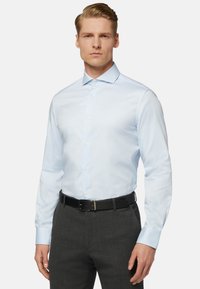 Camicia a maniche lunghe azzurra chiara con colletto a punta, chiusura con bottoni e texture liscia, indossata con pantaloni grigi e una cintura nera.
