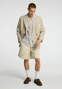 Blazer beige de doble botonadura, camisa de botones a rayas, pantalones cortos crema, zapatos marrones, calcetines blancos. Diseño simple, textura suave y tela ligera.