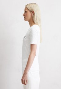 Hvid kortærmet t-shirt med rund halsudskæring og subtil tekstur. Logo trykt på forsiden. Model står i profil.