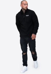 Czarny polar z suwakiem na ćwierć długości, z małym białym logo, połączony z przetartymi czarnymi dżinsami i białymi sneakerami.