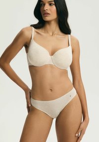 Žena oblečená v béžovom zodpovedajúcom podprsenke a nohavičkách, stojaca s jednou rukou na bokoch, pozerajúca sa do strany na jednoduchom pozadí.