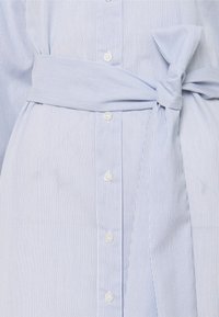 Světle modrá a bílá pruhovaná košile s opaskem uvázaným v pase, zapínání na knoflíky a hladká textilie.