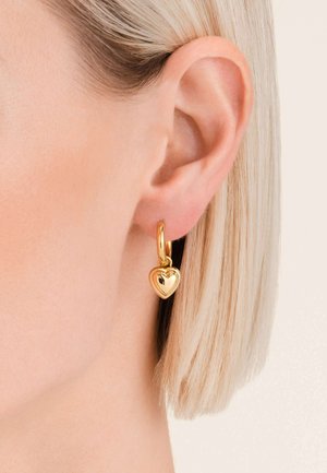 Zlatni naušnicu u obliku srca s poliranim završetkom, obješenu na maloj šarkici. Prikazana na teksturiranom uhu.