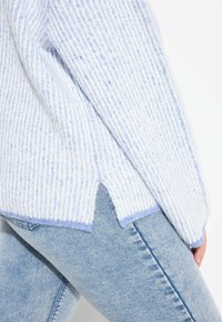 Maglione a righe blu chiaro e bianco con texture a coste, fessure laterali e orlo blu chiaro a contrasto. Abbinato a jeans in denim blu chiaro.