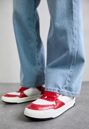 Zapatillas de hombre con detalles en cuero rojo, laterales blancos y suela de goma, combinadas con unos vaqueros de mezclilla sueltos y de ajuste ligero en azul claro.