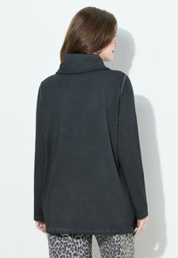 Mujer de espaldas vestida con una camiseta de cuello alto negra de manga larga y pantalones gris con estampado de leopardo, frente a un fondo blanco.