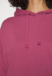 Felpa rosa con cappuccio e lacci con estremità a frange; realizzata in tessuto morbido con una superficie liscia e una tasca frontale.