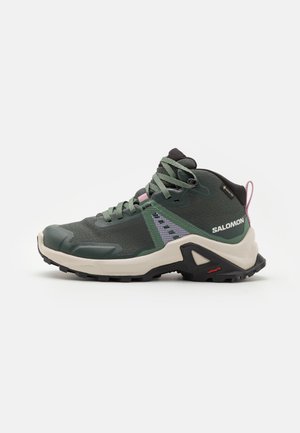 Zapato de senderismo verde con parte superior de malla, acentos en negro y rosa, suela de goma y puntera reforzada con un diseño estampado en el lateral.