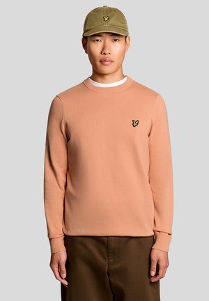 Jonge man die een perzikkleurige trui met ronde hals en een olijfgroene pet draagt, staand tegen een effen lichtgrijze achtergrond.