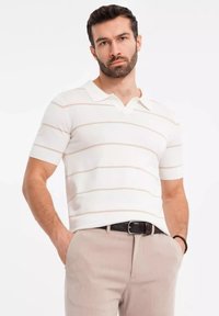 Valkoinen lyhythihainen polo paita vaaleanbeigeillä vaakasuorilla raidoilla, jossa on klassinen kaulus ja teksturoitu kangas. Yhdistetty vaaleanbeigeihin housuihin.