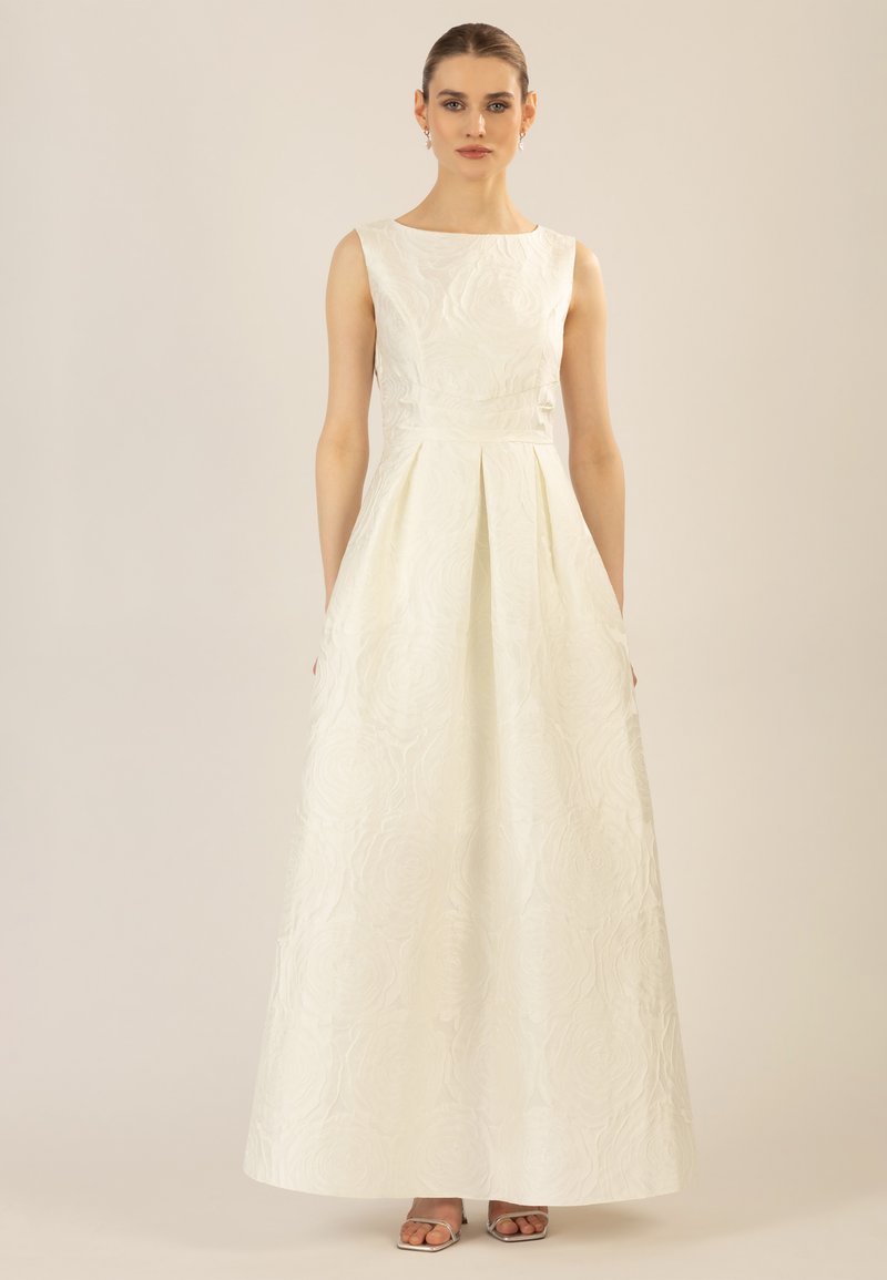 Robe blanche à texture florale avec un corsage ajusté, une jupe plissée évasée et sans manches. Accents argentés sur les sandales. Tissu lisse.