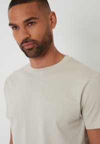 Camiseta de algodón color beige claro con cuello redondo, mangas cortas y un pequeño logo bordado en el lado izquierdo del pecho. Textura suave.