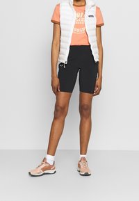 Osoba oblečená v bílé prošívané vestě, korálově oranžovém tričku s potiskem, černých šortkách, bílých ponožkách a korálově oranžových sportovních botách stojící před jednobarevným pozadím.