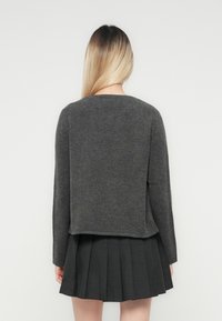Femme avec de longs cheveux blonds portant un pull gris foncé et une jupe plissée noire, se tenant face à un fond blanc uni.