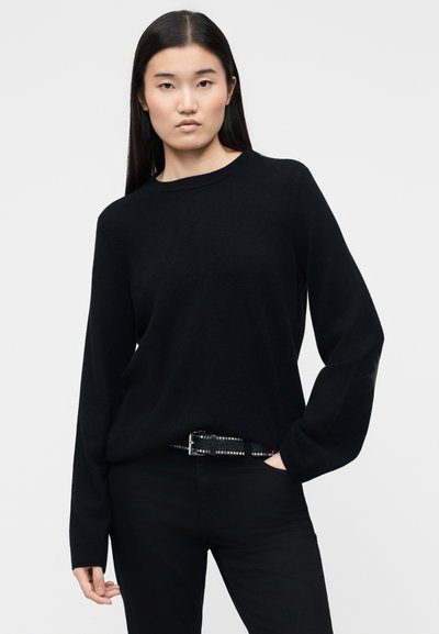 Ασιάτισσα με μακριά μαύρα μαλλιά, φοράει φαρδύ μαύρο πουλόβερ μπα tucked μέσα σε μαύρο παντελόνι με μαύρη ζώνη με τρουκς, στέκεται με το ένα χέρι στην τσέπη.