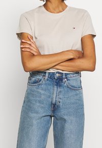 Camiseta de algodón beige con mangas cortas y escote redondo, que presenta un pequeño logo. Combinada con unos vaqueros de cintura alta en azul claro.