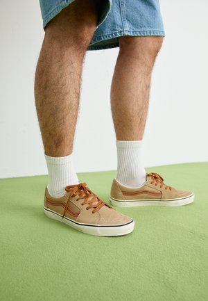 Baskets en suède beige avec des accents marron, semelle en caoutchouc blanc et lacets marron, accompagnées de chaussettes blanches. Fond texturé vert.