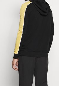 Czarna bluza z kapturem z żółtym panelem na ramionach i białymi akcentami, z kangurzą kieszenią i ściągaczami przy rękawach. Gładki, teksturowany materiał.