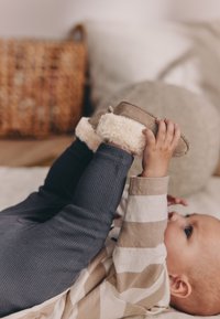 Jasnobrązowe dziecięce kapcie z futerkiem, noszone przez dziecko w paskowanej beżowo-białej koszulce, leżące na fakturowanej powierzchni.