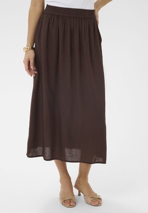 Frau trägt einen braunen Midirock mit elastischem Bund und beigen offenen Absatzschuhen, steht vor einem schlichten weißen Hintergrund.