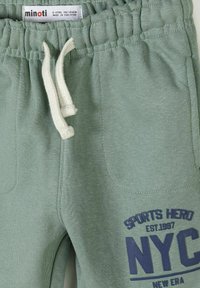 Pantalon de survêtement vert pour enfants avec cordon blanc, taille élastique et l'inscription "SPORTS HERO NYC" en bleu sur la zone de la poche avant.