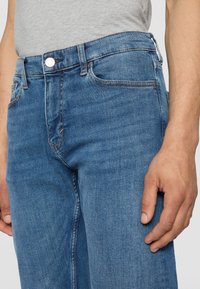 Gros plan d'une personne portant un jean bleu en denim et un t-shirt gris clair, la main droite détendue le long du corps.