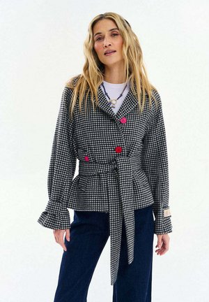 Kobieta o długich blond włosach, ubrana w czarno-białą kurtkę w pepitkę z paskiem i kolorowymi guzikami oraz ciemnoniebieskie spodnie na jednolitym tle.