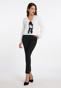 Weiße Strickjacke mit V-Ausschnitt und schwarzem Band zum Binden vorne. Kombiniert mit schwarzen Slim-Fit-Hosen und schwarzen High Heels.
