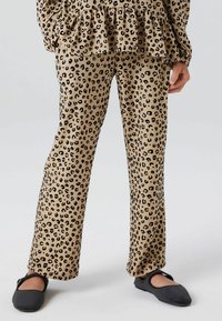 Pantalons en imprimé léopard beige avec des taches noires, fabriqués en tissu doux. Associés à des chaussures plates noires, avec un bout arrondi et une sangle.