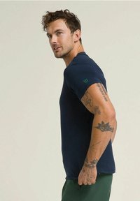 Hombre con cabello rizado que lleva una camisa de manga corta azul marino y pantalones verdes, mostrando tatuajes en su brazo derecho, mirando hacia un lado.