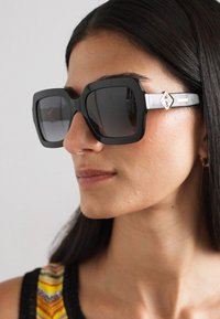 Mustad ümarklaasidega mustad oversize päikeseprillid, millel on ruudukujuline raam ja gradient objektiivid; templis on hõbedane aktsent, millel on geomeetriline disain.