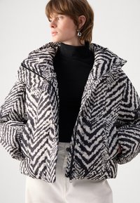 Giacca imbottita con un motivo zigzag bianco e nero, colletto alto e chiusura con zip; tessuto testurizzato, abbinata a un dolcevita nero sotto.