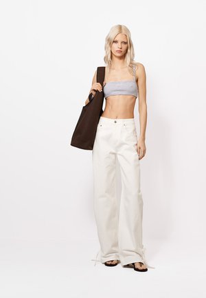 Fiatal nő hosszú szőke hajjal, szürke bandeau felsőt, magas derekú széles fehér nadrágot, fekete szandált visel, és nagy sötétbarna oldaltáskát hord.