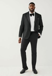 Schwarzer Smoking mit satinierter Revers, weißem plissiertem Hemd und schwarzer Fliege. Abgerundet mit schwarzen Schuhen für einen formellen Auftritt.