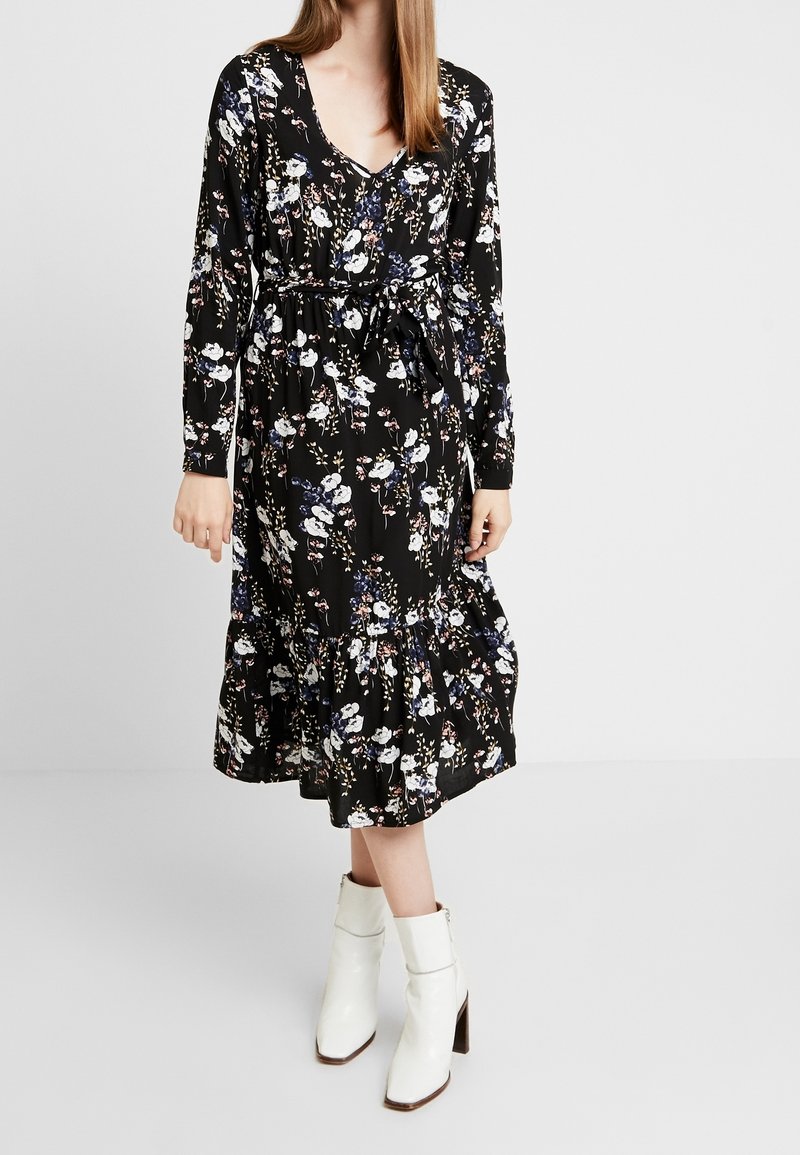 Femme portant une robe midi noire à manches longues à motif floral avec ceinture, associée à des bottines blanches, debout sur un fond uni.