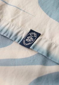 Katoenen stof met blauw-witte cirkelpatronen; voorzien van een marineblauwe label met de tekst "SUPER DRY." Glad van textuur en lichtgewicht uitstraling.