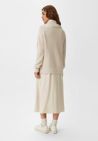 Cremfarbener Rollkragenpullover mit ribbed Textur, kombiniert mit einem fließenden cremfarbenen Midirock. Weiße Sneaker und kniehohe Socken runden den Look ab.
