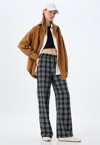 Chemise oversized en daim marron, crop top noir et pantalon large à carreaux noir et blanc, associés à des baskets blanches, le tout sur un fond clair.