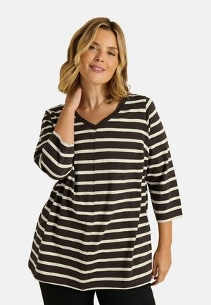 Woman with shoulder-length blonde hair wearing black and white horizontal striped three-quarter sleeve top, smiling with one hand touching her neck.
