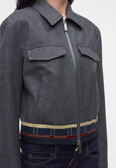 Femme portant une veste grise zippée avec deux poches poitrine et une bordure décorative à carreaux et ruban à la taille.