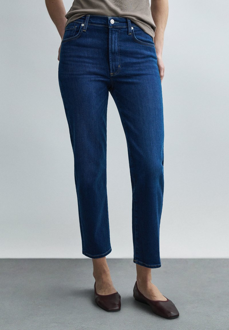 Kék farmernadrág magas derékkal, egyenes szárú kialakítással és öt zsebbel. Látható kopás vagy díszítés nélkül, sötét balerinacipővel párosítva.