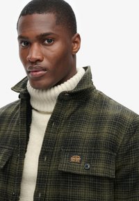 Chaqueta de camisa a cuadros verdes con detalles negros, que cuenta con un bolsillo con botón y un cuello de tortuga de canalé crema debajo.
