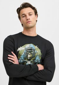 Felpa nera con una grafica di una scimmia in un casco blu che guida una motocicletta, con uno sfondo testurizzato e sfumato in verde e bianco.