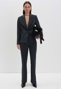 Giacca e pantaloni su misura grigio scuro con una texture liscia, blaser abbottonato e tacchi affilati neri. Il modello tiene una borsa nera con texture.