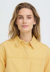 Jeune femme aux yeux bleus et cheveux blonds détachés, portant une chemise jaune boutonnée, face à la caméra avec une expression neutre.