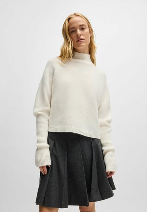 Cremfarbener Strickpullover mit hohem Kragen und langen Ärmeln; kombiniert mit einem dunkelgrauen, gefältelten Rock. Weicher Stoff, verkürzter Schnitt und eng anliegende Bündchen.
