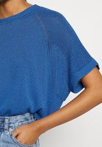 Modrý pletený top s krátkými rukávy, kulatým výstřihem a jemnými rozparky na švech ramen. Nošeno s džínovými šortkami s vysokým pasem.