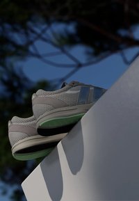 Šedé síťované sportovní boty s zelenými akcenty, které mají texturovaný povrch a horizontální modré pruhy po stranách, umístěné na bílé ploše.